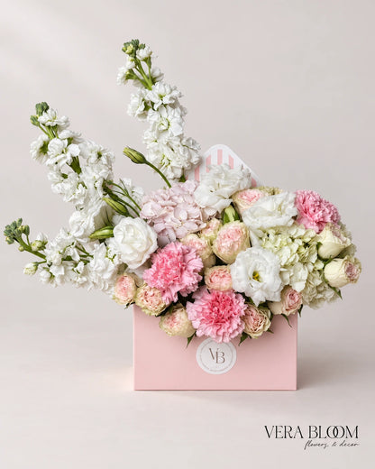 Pastel Letter flower arrangement in a pink envelope box with white, blush and pale green blooms, front view.